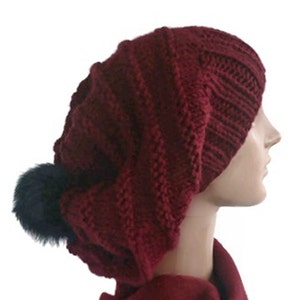 Peut inclure: Un bonnet en tricot bordeaux avec un pompon en fausse fourrure noire. Le bonnet est ample et présente un motif en tricot torsadé.