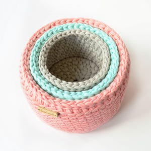 May include: A set of four round, nested baskets in shades of pink, teal, and gray. The baskets are made of a thick, woven material and have a handmade tag attached to the largest basket. The baskets are arranged in a circular pattern.