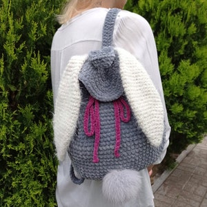 May include: A grey and white crocheted backpack with pink straps and a fluffy white tail. The backpack is shaped like a bunny with long ears.