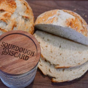 May include: A wooden jar with the text "Sourdough Discard" on the lid, next to a loaf of sourdough bread sliced into four pieces.