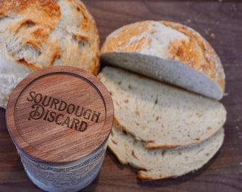 May include: A wooden jar with the text "Sourdough Discard" on the lid, next to a loaf of sourdough bread sliced into four pieces.
