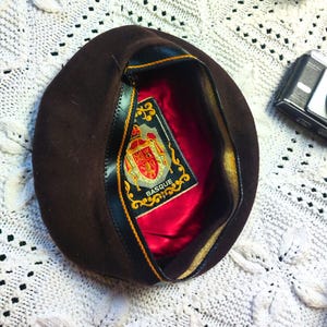 May include: A brown beret with a red satin lining and a gold-trimmed label that reads "Basque." The beret is set on a white, textured surface, with a small camera and several small plants nearby.