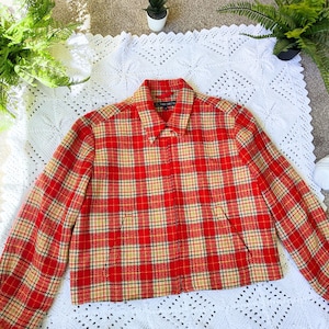 Puede incluir: Chaqueta a cuadros rojos y beige con cuello y mangas largas. La chaqueta está hecha de tela tejida y tiene un corte corto. La chaqueta se exhibe sobre una manta blanca de ganchillo.