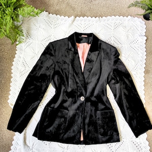 Puede incluir: Un blazer de terciopelo negro con un solo botón y dos bolsillos delanteros. El blazer tiene solapas con muescas y un forro rosa claro. La prenda se muestra sobre una superficie blanca estampada.
