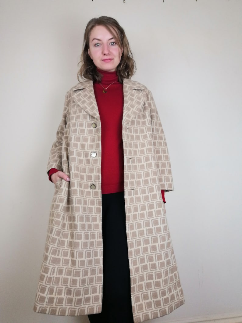 Puede incluir: Un abrigo largo beige con un patr&oacute;n geom&eacute;trico. El abrigo tiene cierre de botones y bolsillos. La modelo lleva un su&eacute;ter rojo de cuello alto y pantalones negros.
