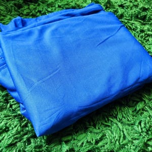 Può includere: Un tessuto blu reale piegato è appoggiato su una superficie verde e pelosa. Una fotocamera vintage è sullo sfondo. La trama liscia del tessuto contrasta con lo sfondo strutturato, creando una composizione visivamente accattivante.