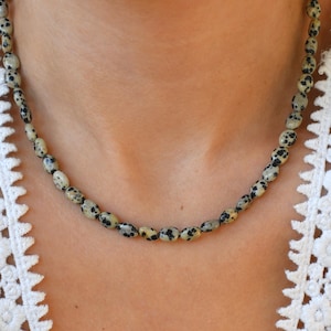May include: A beaded necklace featuring oval-shaped beads in a speckled pattern of black and cream. The necklace is worn around the neck, with the beads resting against the skin. The background is a white lace garment.