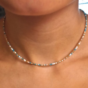 May include: A delicate beaded necklace with small, colorful beads in shades of white, blue, and gold. The necklace is made of tiny beads strung together on a thin chain.