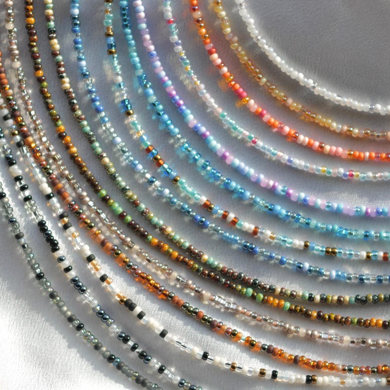 May include: A collection of 20 different beaded necklaces in various colours, including white, black, brown, blue, and pink. The necklaces are made with small, uniform beads and are arranged in a semi-circle pattern.
