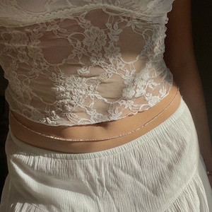 Puede incluir: Un top de encaje blanco con una delicada cadena de cuerpo plateada. La cadena se lleva alrededor de la cintura y es visible contra la tela blanca.