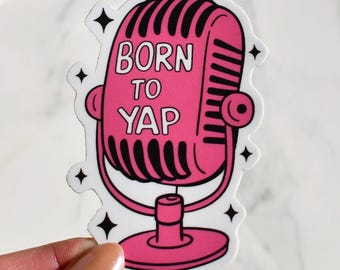 Può includere: Un adesivo rosa con un design a microfono retrò e la scritta bianca "BORN TO YAP". Il microfono è delineato in nero e ha quattro stelle nere intorno. L'adesivo è tenuto da una mano.