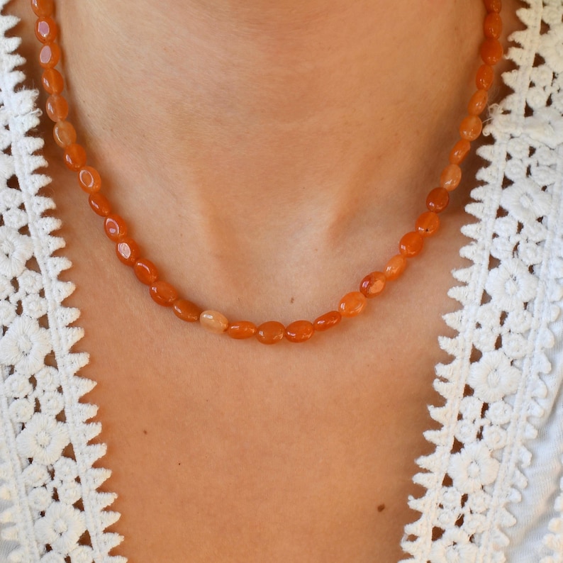 May include: A necklace composed of oval-shaped, orange-colored beads. The beads are strung together to form a necklace. The necklace is worn against a white lace top.