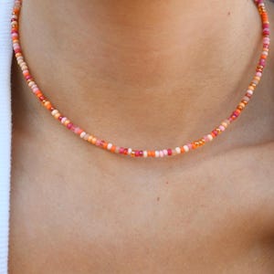 May include: A beaded choker necklace featuring a repeating pattern of small, round glass beads in various shades of orange, pink, coral, and peach. The necklace is strung on a thin cord and sits closely around the neck, displaying a vibrant colour palette.