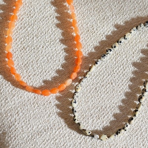 May include: Two beaded necklaces. One necklace features orange oval beads. The other necklace has off-white beads with black spots. Both necklaces are displayed on a textured, off-white surface.