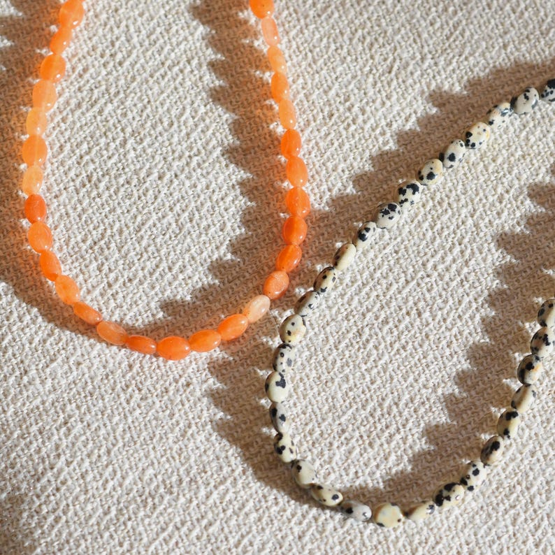 May include: Two beaded necklaces. One necklace features orange oval beads. The other necklace has off-white beads with black spots. Both necklaces are displayed on a textured, off-white surface.