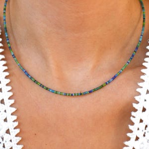May include: A finely beaded necklace displaying a gradient of blue and green beads. The necklace is set against a white backdrop, accentuating the vibrant colours of the beads. The necklace is a simple, elegant accessory.
