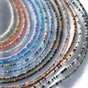 May include: A collection of colourful beaded necklaces in various shades of blue, orange, pink, brown, and white. The necklaces are arranged in a fan shape on a white surface.