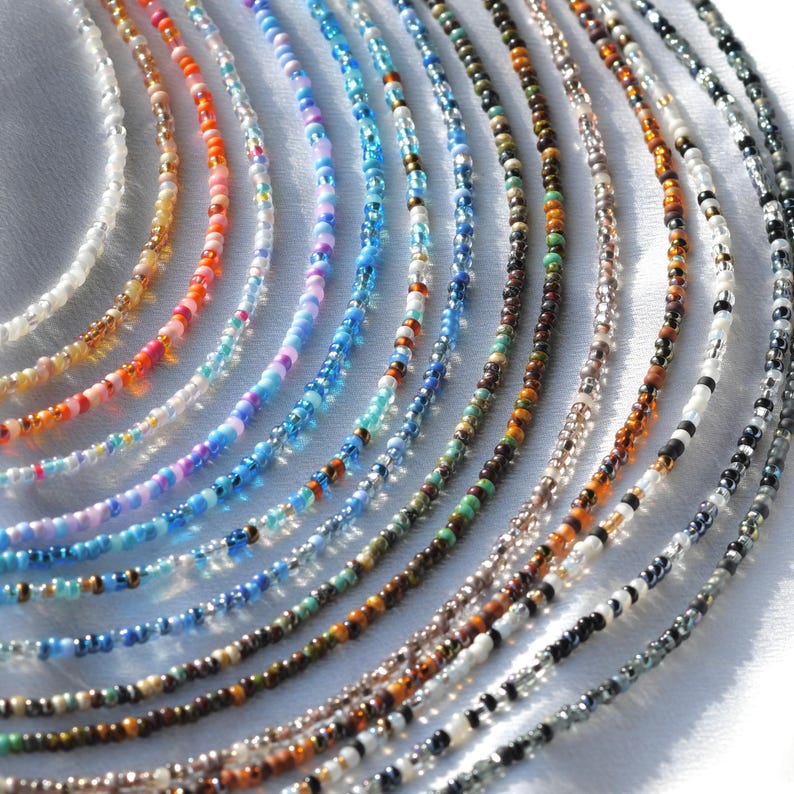 May include: A collection of colourful beaded necklaces in various shades of blue, orange, pink, brown, and white. The necklaces are arranged in a fan shape on a white surface.