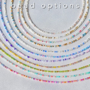 May include: A collection of eleven circular beaded necklaces arranged in a fan pattern, each featuring distinct color combinations labeled as moonstone, alabaster, spring flowers, ocean coast, snowflake, cotton candy, amethyst, spring suns, meadows, mermaid beach, and sunset, with the text bead options displayed at the top.