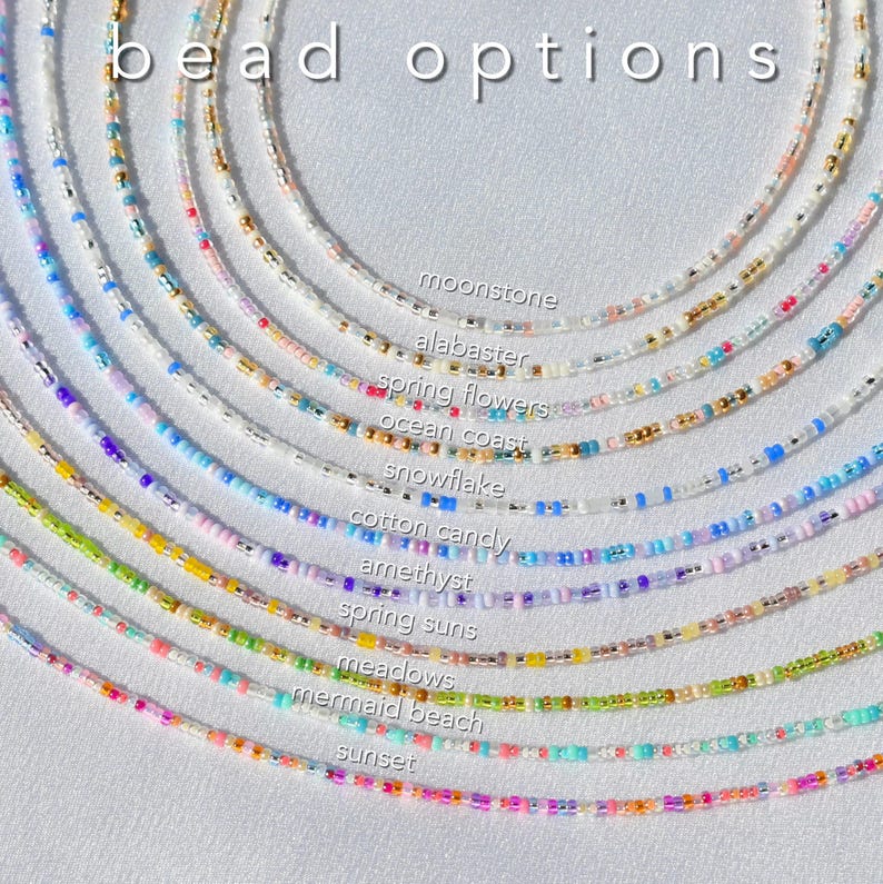 May include: A collection of eleven circular beaded necklaces arranged in a fan pattern, each featuring distinct color combinations labeled as moonstone, alabaster, spring flowers, ocean coast, snowflake, cotton candy, amethyst, spring suns, meadows, mermaid beach, and sunset, with the text bead options displayed at the top.