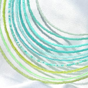 Op de afbeelding: Een verzameling kralenkettingen in groen- en blauwtinten. De kettingen zijn gemaakt van kleine, ronde kralen en zijn in een gebogen patroon gerangschikt. De kleuren variëren van lichtgroen tot turquoise en aqua, waardoor een gradiënteffect ontstaat. De achtergrond is een witte, getextureerde stof.