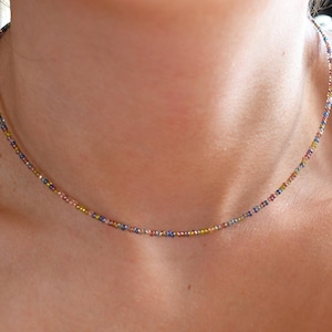 Peut inclure: Collier délicat composé de petites perles multicolores roses, bleues, jaunes et dorées. Le collier est porté autour du cou, mettant en valeur le design complexe et les couleurs vives. Un bijou simple et élégant.