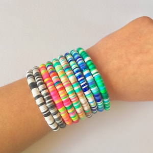 以下が含まれることがあります： 小さな丸いビーズで作られたカラフルなブレスレットのスタック。ブレスレットは人の手首に重ねてあり、白、黒、ピンク、青、緑、黄など、さまざまなパターンと色があります。