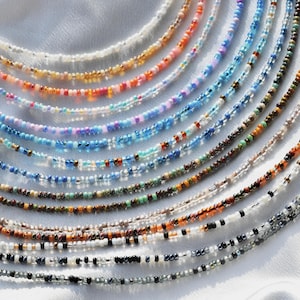 May include: A collection of colourful beaded necklaces in various shades of blue, brown, orange, white, and black. The necklaces are arranged in a fan shape on a white surface.