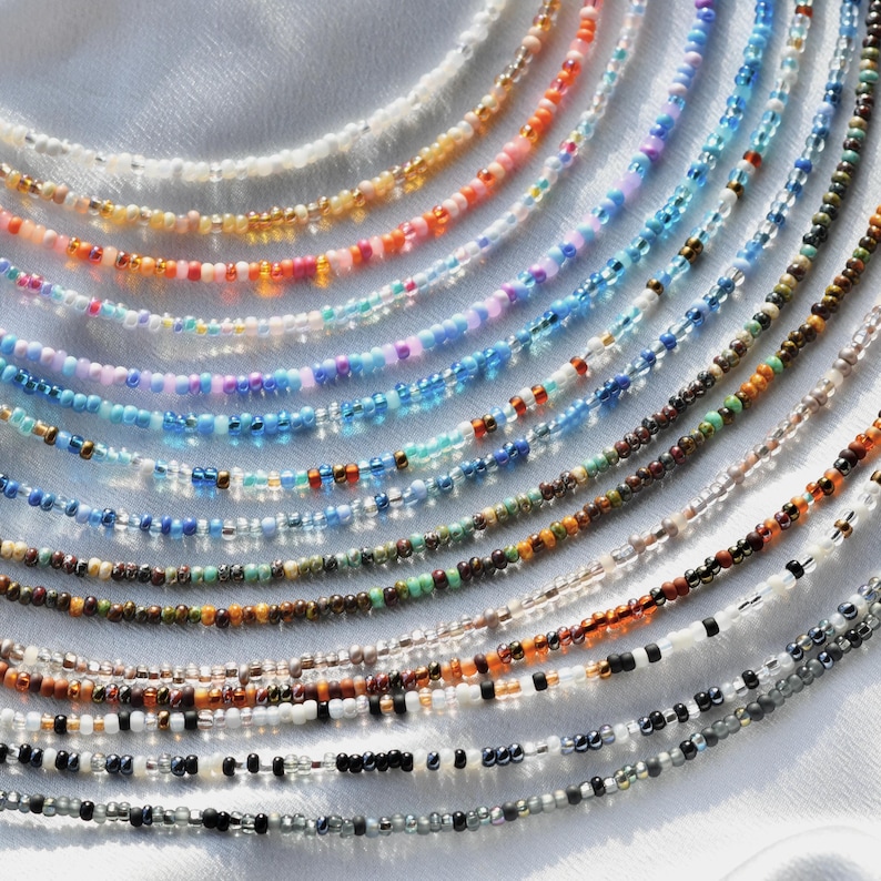 May include: A collection of colourful beaded necklaces in various shades of blue, brown, orange, white, and black. The necklaces are arranged in a fan shape on a white surface.