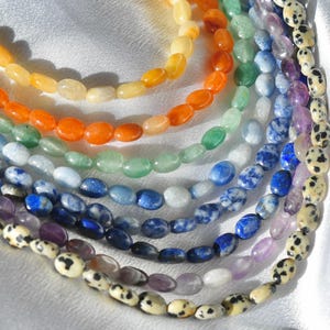 May include: A collection of gemstone bead necklaces in a rainbow of colors. The beads are oval-shaped and strung together, showcasing a variety of hues from yellow and orange to green, blue, and purple. Some beads have speckled patterns.