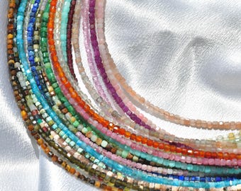 Könnte beinhalten: Eine Sammlung zarter Edelstein-Halsketten in verschiedenen Farben, darunter Blau, Grün, Orange, Rosa und Lila. Jede Halskette besteht aus kleinen, quadratisch geschliffenen Perlen, wodurch eine strukturierte und farbenfrohe Darstellung entsteht. Die Halsketten sind auf einer weißen, seidigen Oberfläche angeordnet.