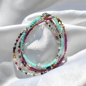 Può includere: Un set di quattro braccialetti di perline nei toni del turchese, viola e pietre preziose multicolori. I braccialetti sono infilati su catene d'argento e chiusi con una chiusura d'argento. Esposti su un tessuto bianco e setoso.