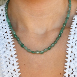 May include: A green beaded necklace with oval-shaped beads. The necklace is worn against a white lace top. The beads are a mix of light and dark green, creating a natural, earthy look. The necklace appears to be a single strand.