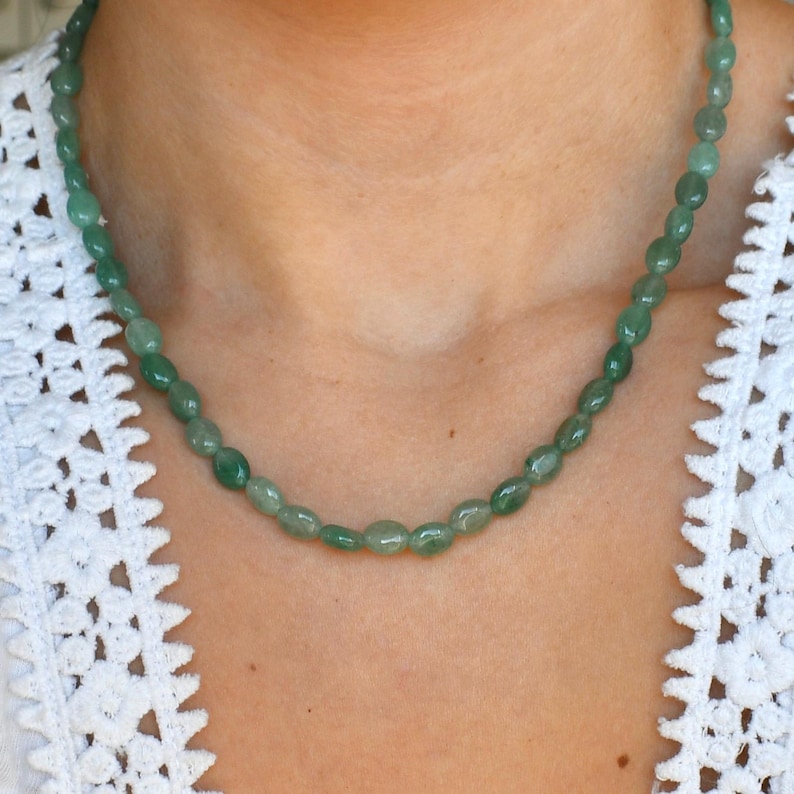May include: A green beaded necklace with oval-shaped beads. The necklace is worn against a white lace top. The beads are a mix of light and dark green, creating a natural, earthy look. The necklace appears to be a single strand.
