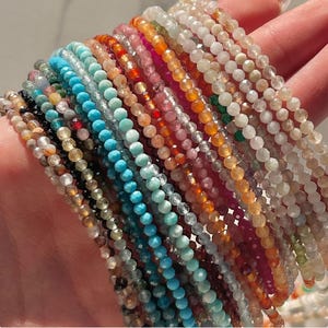 May include: A collection of 20 strands of gemstone beads in various colors, including turquoise, pink, green, and brown. The beads are small and uniform in size, and they are strung on clear thread.