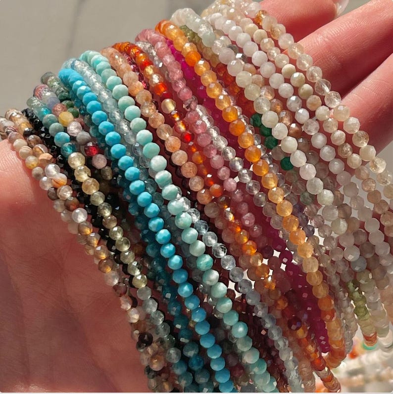 May include: A collection of 20 strands of gemstone beads in various colors, including turquoise, pink, green, and brown. The beads are small and uniform in size, and they are strung on clear thread.