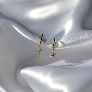 May include: A pair of gold hoop earrings with a dangling star charm. The star has a small purple gemstone in the center.