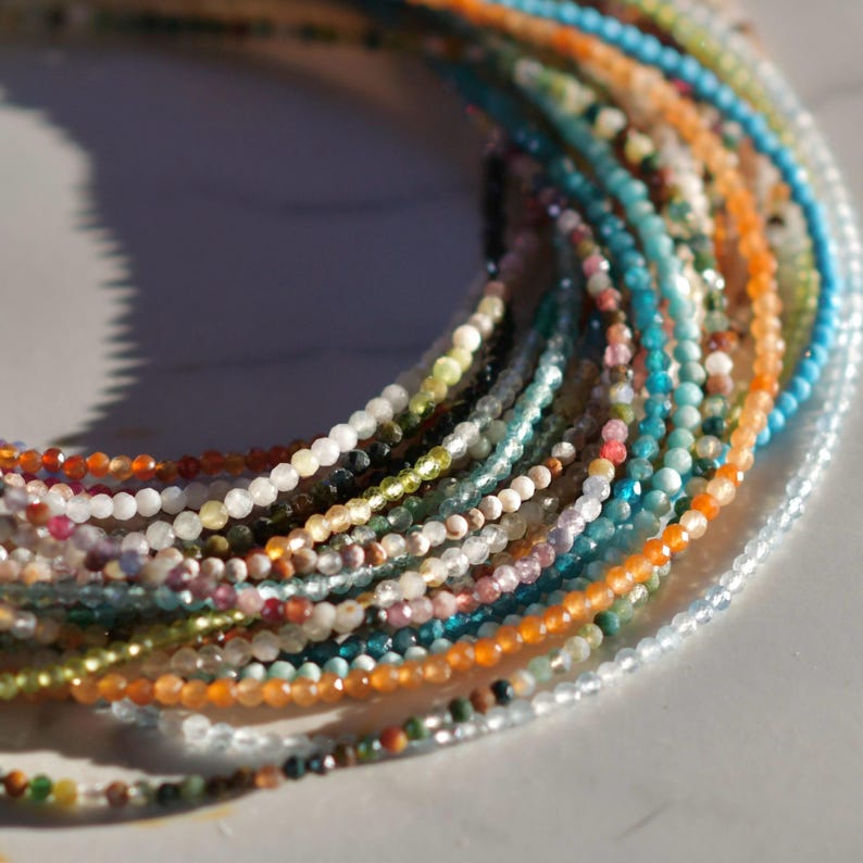 May include: A close-up of a collection of colorful gemstone beads strung on thin cords. The beads are arranged in a circular pattern, creating a layered effect. The colors of the beads include blue, green, orange, red, and white.