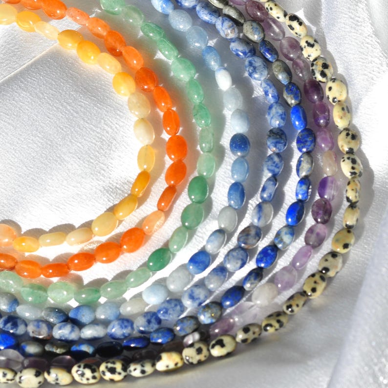 May include: A collection of gemstone bead strands in a rainbow of colors. The beads are oval-shaped and arranged in a curved pattern, showcasing a spectrum from yellow and orange to green, blue, and purple, with a final strand of spotted beads.