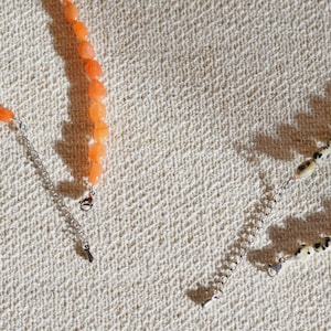 May include: Close-up of two necklaces. One necklace features orange gemstone beads, while the other has cream-colored beads with black spots. Both necklaces have silver-colored chain extensions and clasps. The background is a textured, off-white fabric.