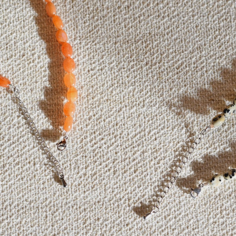 May include: Close-up of two necklaces. One necklace features orange gemstone beads, while the other has cream-colored beads with black spots. Both necklaces have silver-colored chain extensions and clasps. The background is a textured, off-white fabric.