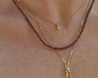 May include: Three gold necklaces layered on a person's neck. The top necklace has a small, round, clear gemstone pendant. The middle necklace is a thin gold chain. The bottom necklace is made of small, dark red beads.