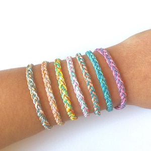 May include: A set of eight colorful braided bracelets in shades of pink, blue, green, and yellow. The bracelets are made of thin, woven cords and are stacked on a person's wrist.