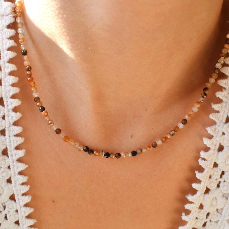 May include: A delicate necklace made of small, multi-colored gemstone beads. The beads are a mix of brown, tan, and black, and they are strung on a thin silver chain.