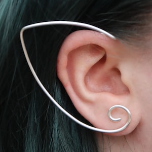 Może przedstawiać: Srebrny nausznik w kształcie uszu elfa. Nausznik zakrzywia się wokół ucha i ma spiralny wzór na dole. Ucho osoby i jej niebiesko-zielone włosy są widoczne w tle.