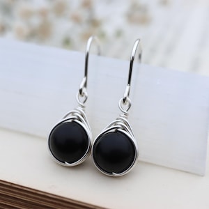 Puede incluir: Un par de pendientes colgantes plateados con piedras negras redondas. Las piedras están engarzadas en un diseño envuelto en alambre. Los pendientes se muestran sobre una superficie blanca con un fondo suave y borroso.