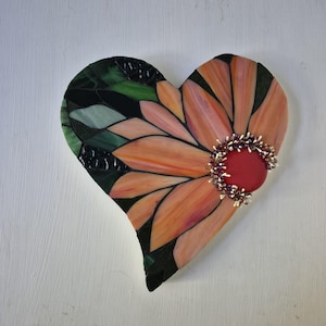 May include: A heart-shaped stained glass mosaic featuring peach-coloured petals radiating from a central solid red circular cabochon. The outer edges are composed of dark green and black glass pieces, with small decorative beads surrounding the centre.