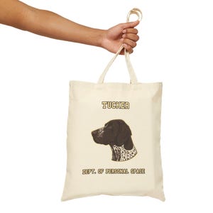 May include: Cream-colored tote bag featuring a brown and white dog illustration with the name "Tucker" above and "DEPT. OF PERSONAL SPACE" below. The bag is held by a person's hand, showcasing its design and handle.