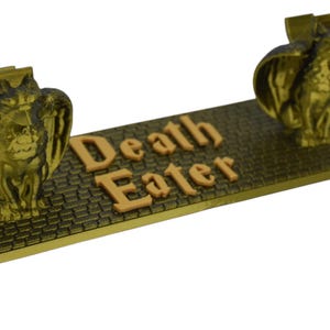 Könnte beinhalten: Ein goldfarbener Metall-Buchstütze mit zwei Gargoyle-Figuren auf jeder Seite. Die Buchstütze hat ein schwarzes, ziegelartiges Muster und die Worte "Death Eater" in orangefarbener Schrift.