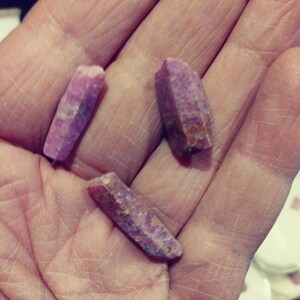 May include: Three pink and purple crystal points, with a rough, natural texture, are held in a hand.